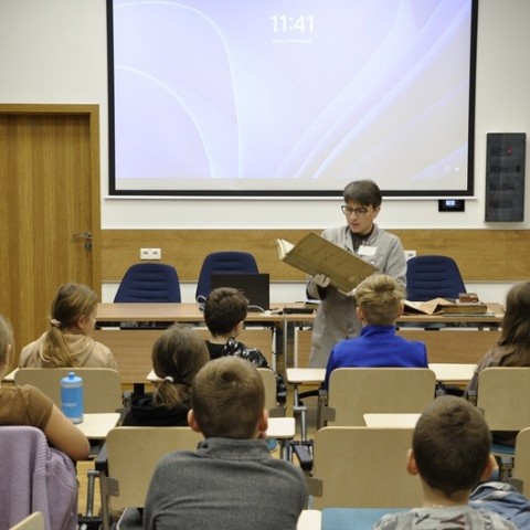 Powiększ obraz: grupa dzieci siedzi frontem do archiwistki pokazującej akta