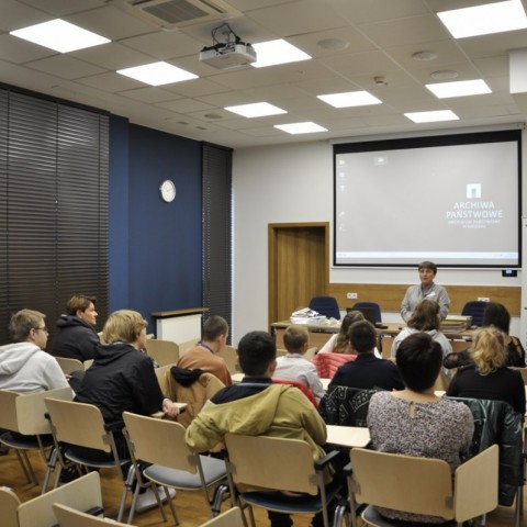 Powiększ obraz: grupa dzieci siedząca na krzesełkach w sali konferencyjnej w kierunku archiwistki